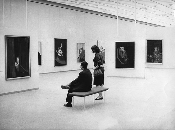 Black-and white-photograph of two people with their backs to the camera, looking at the Francis Bacon paintings hung before them in the gallery space.