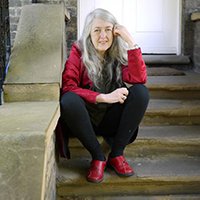 Professor Dame Mary Beard FBA shown sitting on some steps and smiling to the camera, wearing a red coat and red shoes.