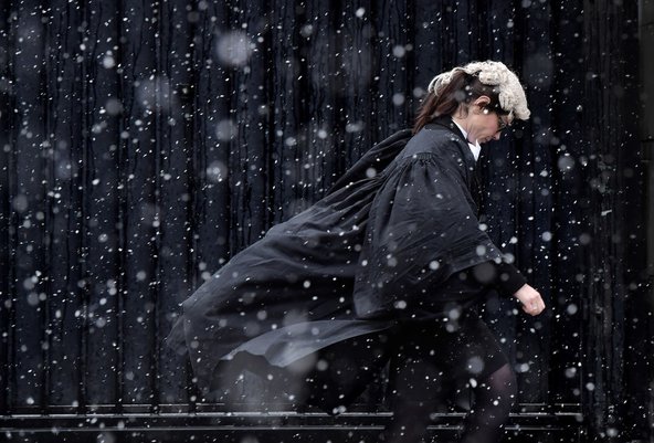 A barrister wearing her wig and gown walks purposefully through falling snow with her head down.