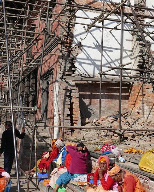 People sit outside scaffolding and a collapsed building in Kathmandu
