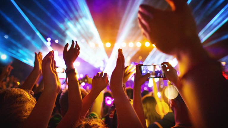 People at a concert, dancing with their hands raised as strobe lights flash above them. One concert goer is filming on their mobile phone.