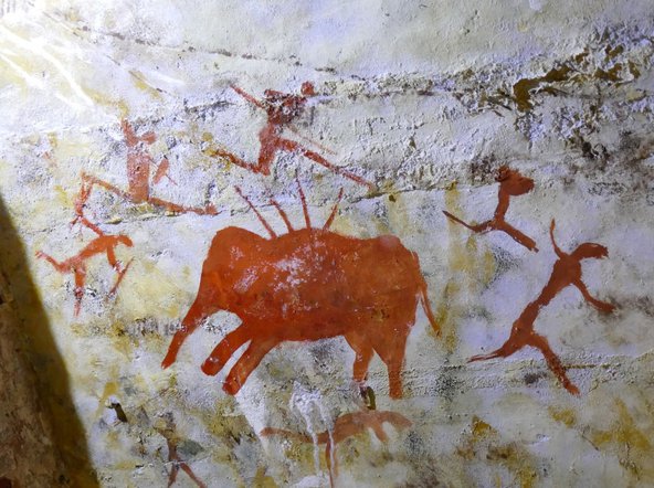 Colourful photograph of a cave painting showing an orange elephant with spears sticking out of its back and orange stick men with weapons around it.