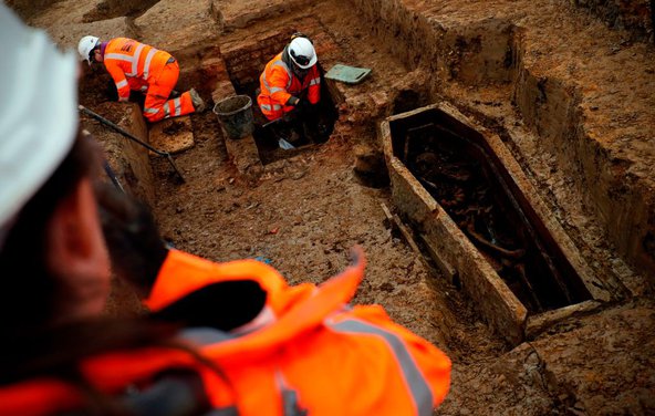 Photograph showing archeologists in orange suits and safety helmets excavating a coffin.