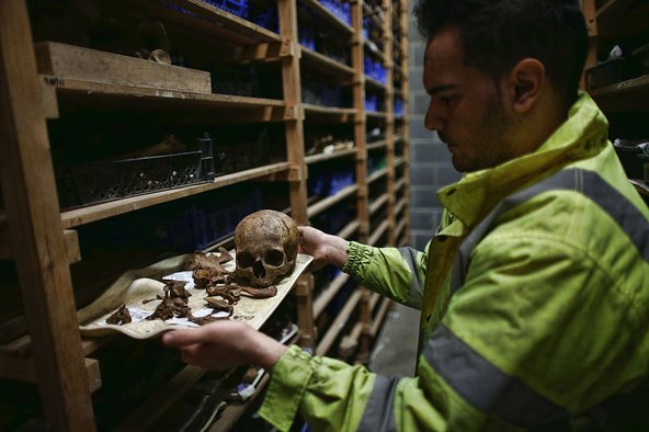 Photograph of a professional in a yellow reflective suit next to a wooden shelf with human remains including a skull and bones.